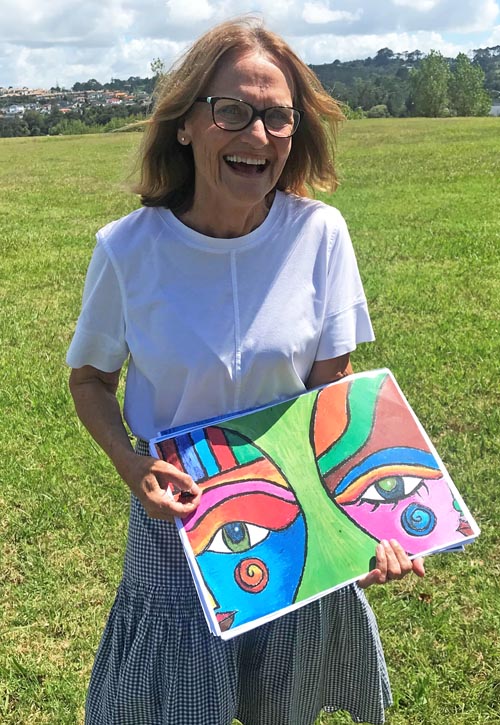 Gwen Taylor is standing in a grassy space with a smile on her face and holding an artwork in her hands