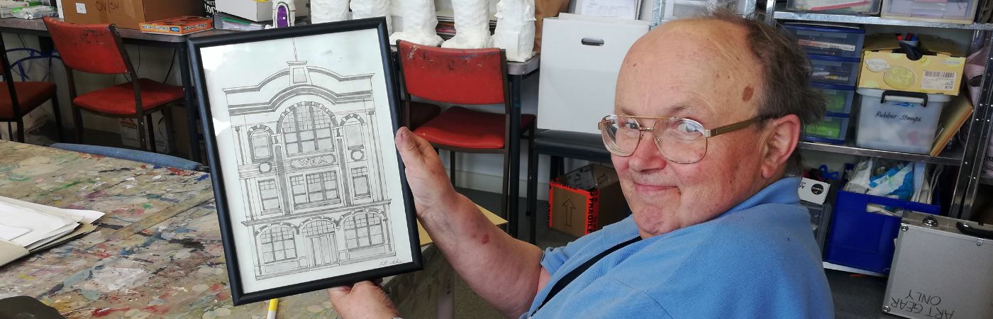 A man wearing glasses and a light blue shirt sits in a creative studio, holding up a framed architectural drawing of a historic building. The room is filled with art supplies, sculptures, and colourful artwork on the walls.