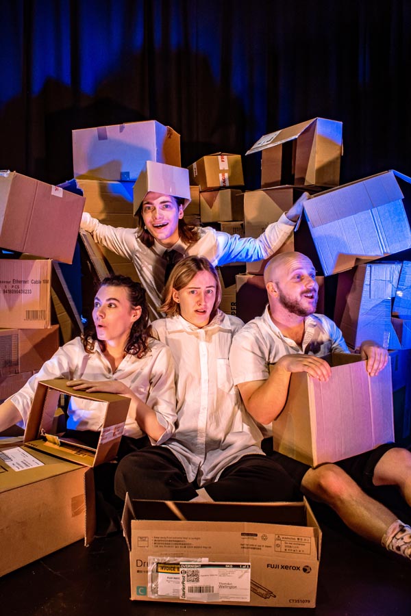 Four cast members sit on the stage surround by a pile of cardboard cartons