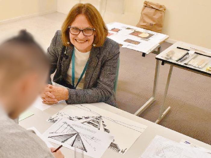 Gwen Taylor is sitting at a desk with artwork on it. She is smiling and looking at the blurred face of a man, whom we cannot identify.