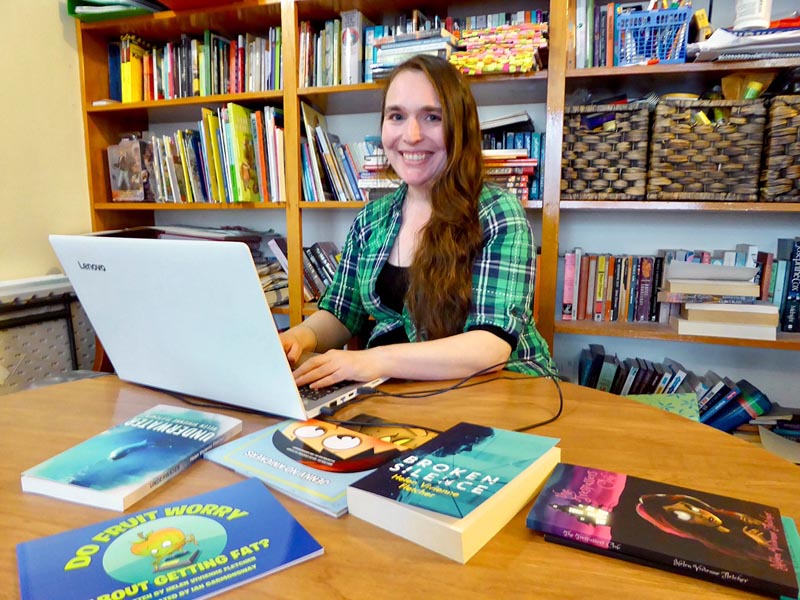Helen Vivienne Fletcher sits at a desk writing on a laptop. She has long brown hair and is wearing a green checked shirt. The table is covered with books.