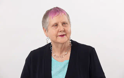 Robyn Hunt ONZM is photographed against a blank white wall. She wears a black cardigan, teal top, silvery chain necklace and earrings and her signature pink fringe.