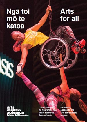 Two dancers, Rodney Bell and Brydie Colquhoun, perform Hurihuri. Rodney is in a wheelchair in the air and his right hand reaches down to hold Brydie's foot. Her body is extended and she holds on to the wheelchair with her left hand.