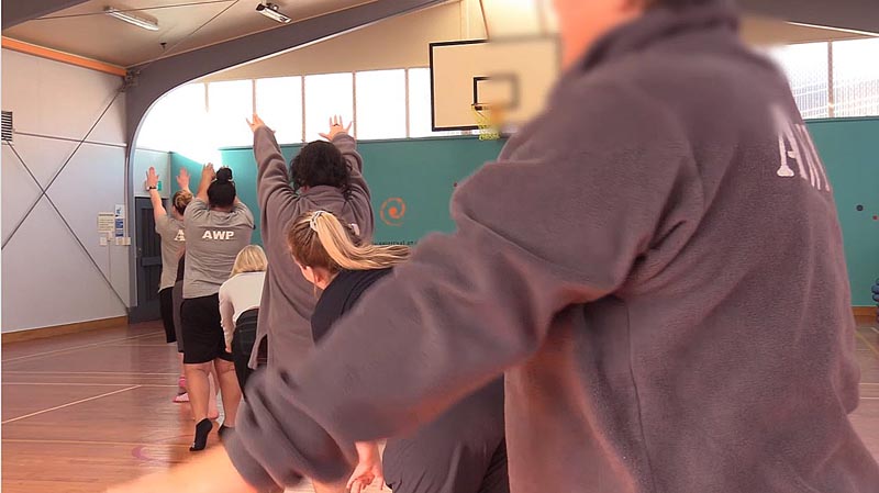 Kristie Mortimer teaches dance at Arohata Women's Prison. The image is of women in a line, wearing brown sweaters with the words AWP on their backs. 