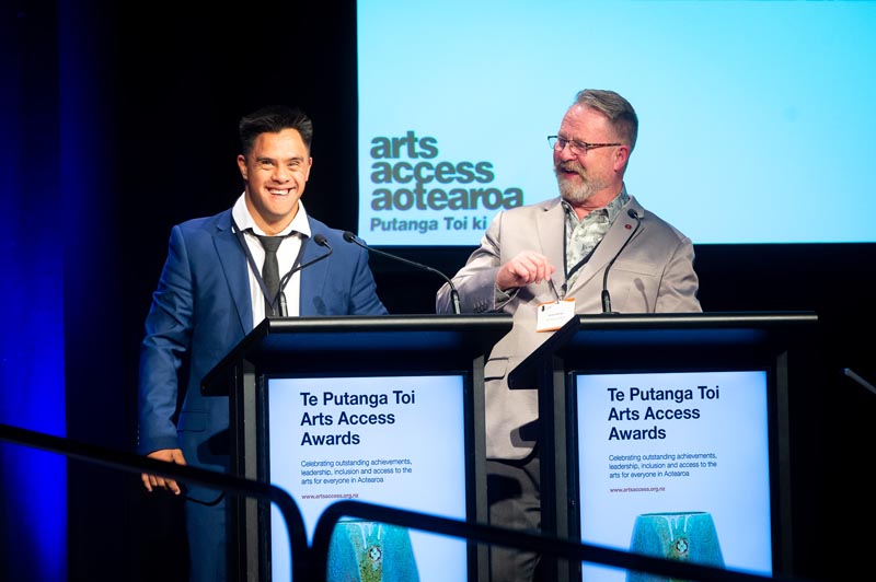Jacob Dombroski and Arts Access Aotearoa Executive Director Richard Benge hosting Te Putanga Toi Arts Access Awards 2021 stand behind two lecterns at Te Papa. They are laughing and behind them is the orange logo of Arts Access Aotearoa