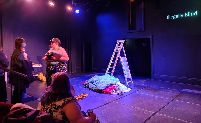 Susan, a blind person holding an armful of braille and text programmes greets audience members as they enter the theatre. On the stage behind them is a ladder and a colourful pile of laundry. White captions on the black wall behind read Illegally blind