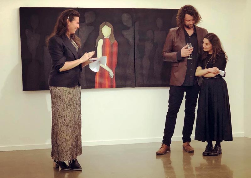 Exhibition curator Meredith Robertshawe, Richard Harkness, and Maisie Chilton standing in front of Maisie's painting 'Ghosts' at the opening of Where does it hurt?