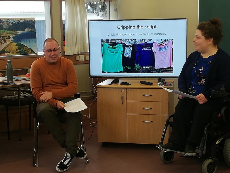 Stace Robertson and Erin Gough lead a disability workshop for Arts Access Aotearoa staff. They are on either side of a television screen, which has an image with the text 'Cripping the script' and a washing line of colourful tee-shirts hanging on it