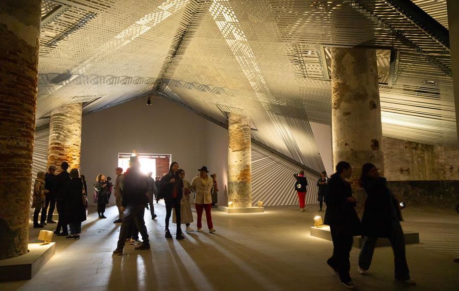 Installation art titled Takapau (2022) by Mataaho Collective, installed at the 2024 Venice Biennale. People walking underneath a woven roof with 4 large pillars in dim light