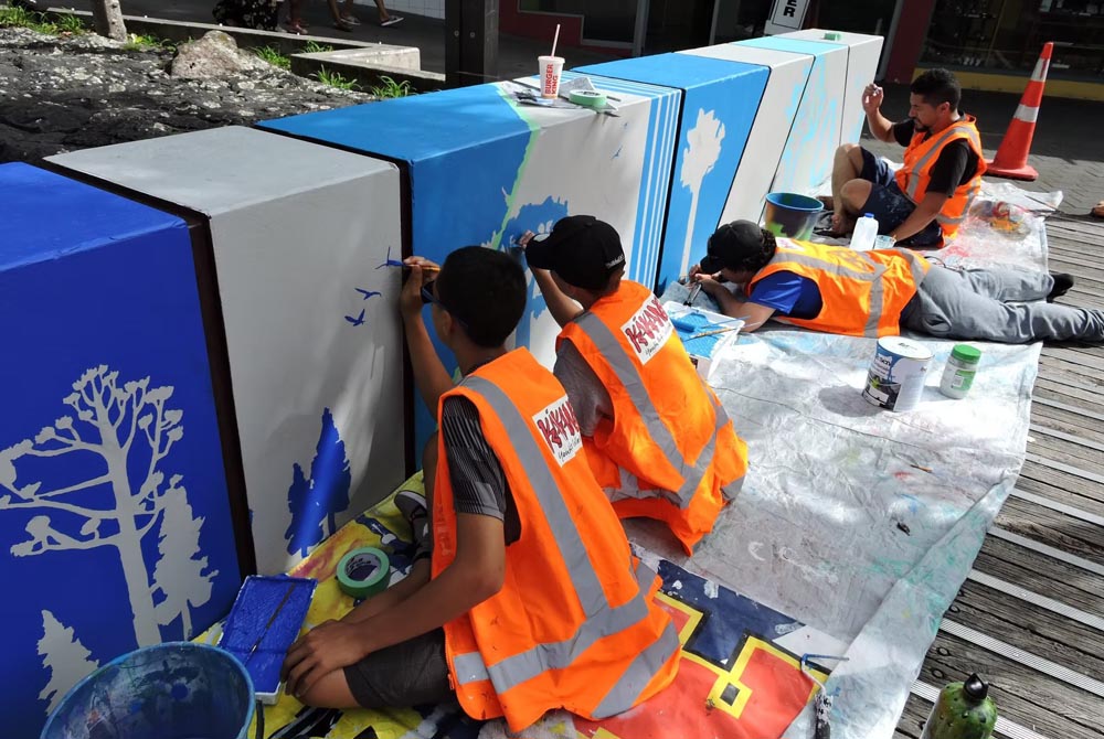 Four Kākano artists, wearing orange hi-vis vests, painting a mural on a wall in the community 