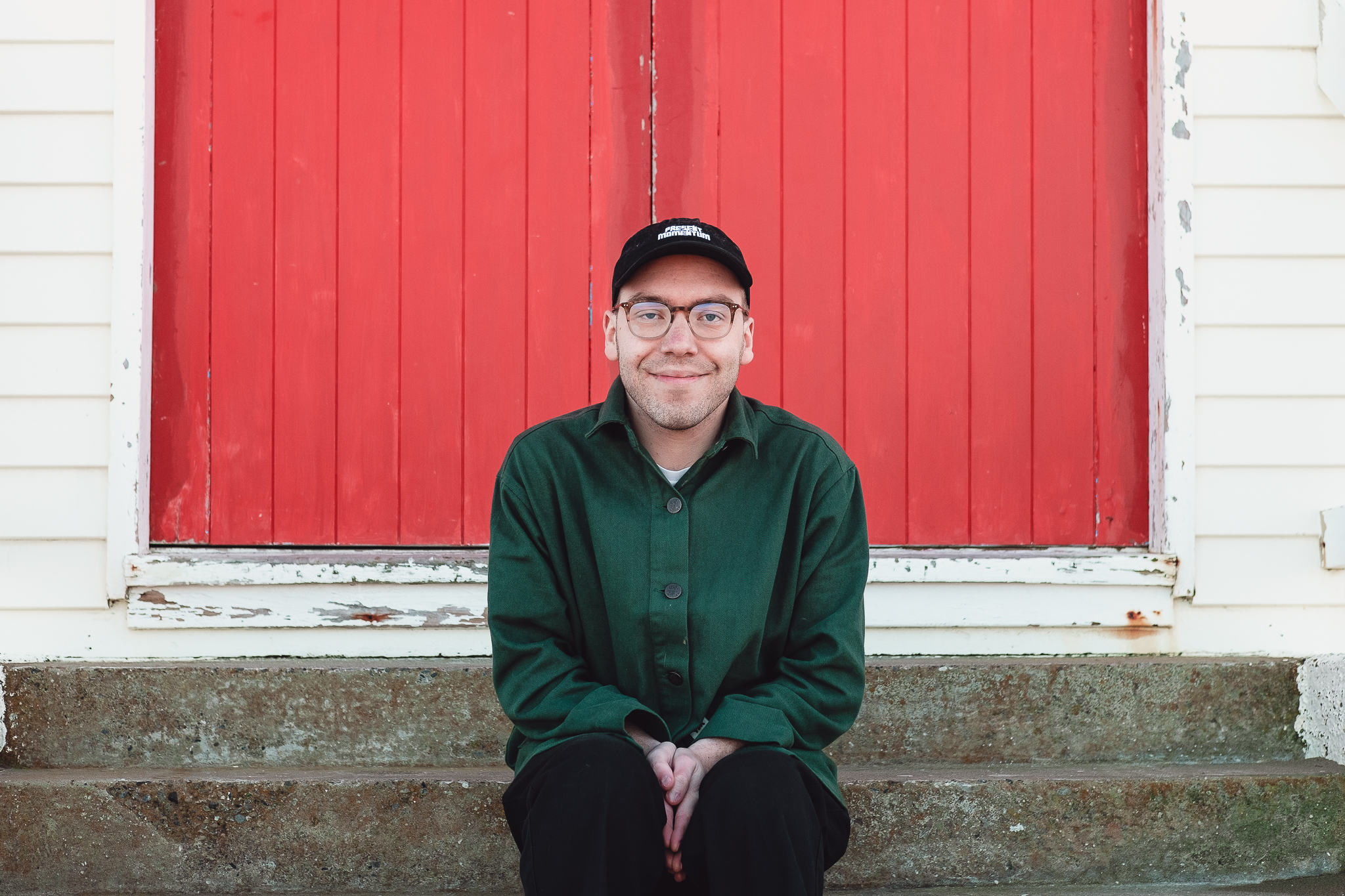 Jesse Austin-Stewart sits on concrete steps in front of a red-painted wooden door.
