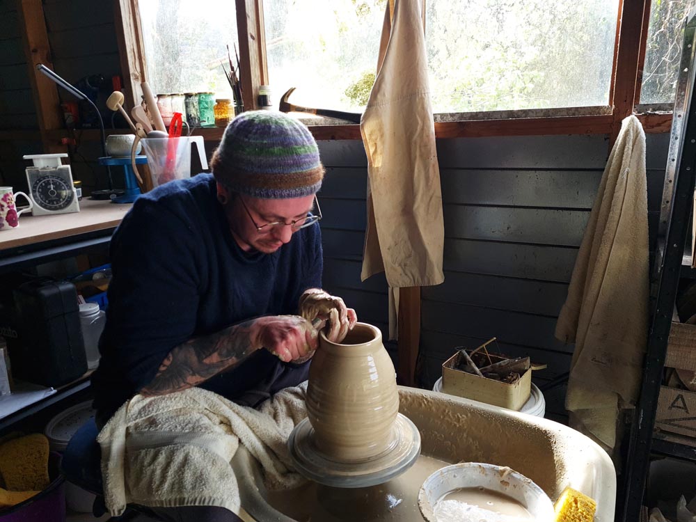 Stace sits at a pottery wheel throwing a large vase. 