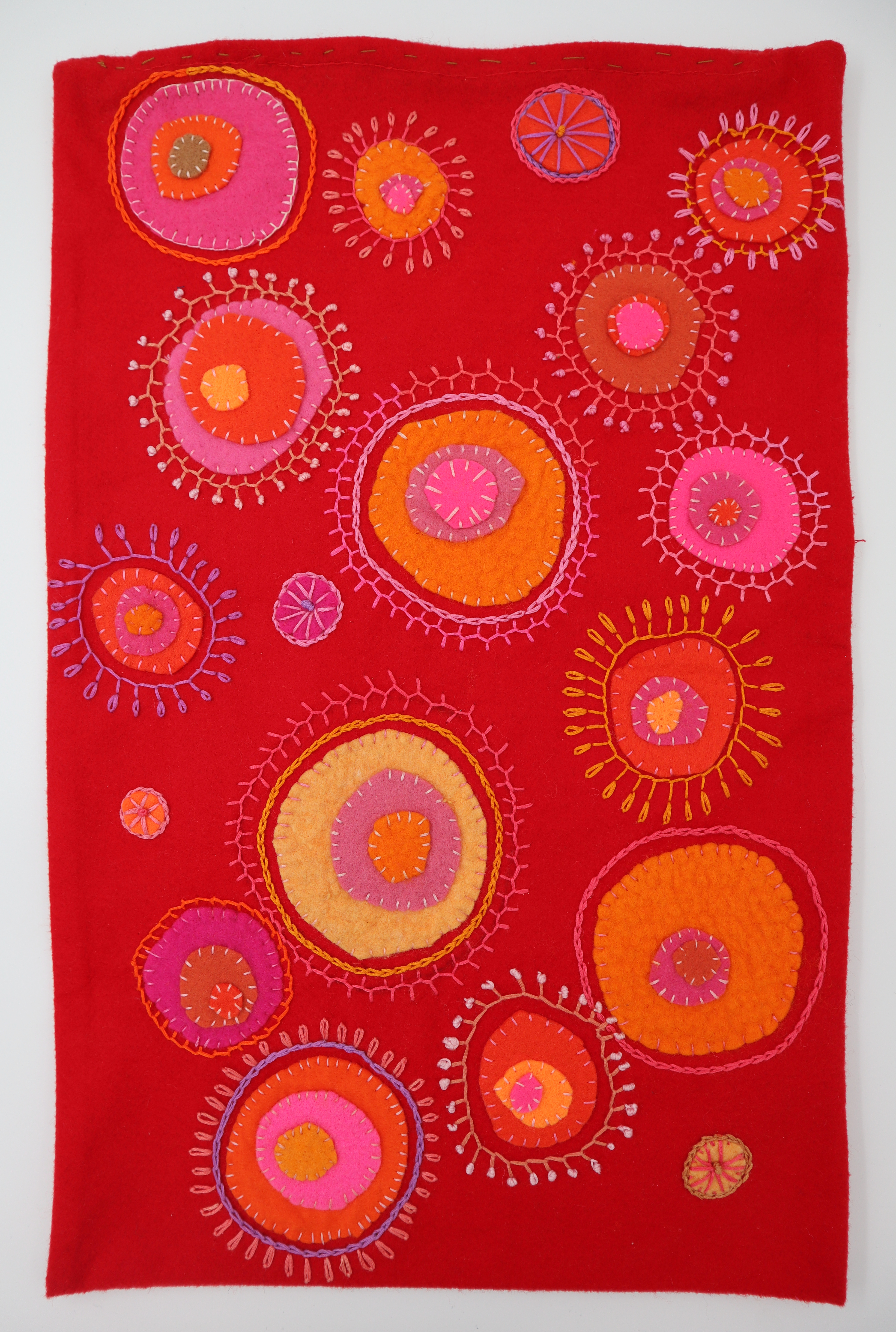 An embroidered cushion cover. Brightly coloured stitching in circular designs on a red felt background