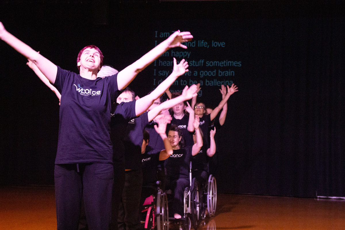 A row of dancers lined up behind one another stand and sit with arms outstretched in a v shape skyward.