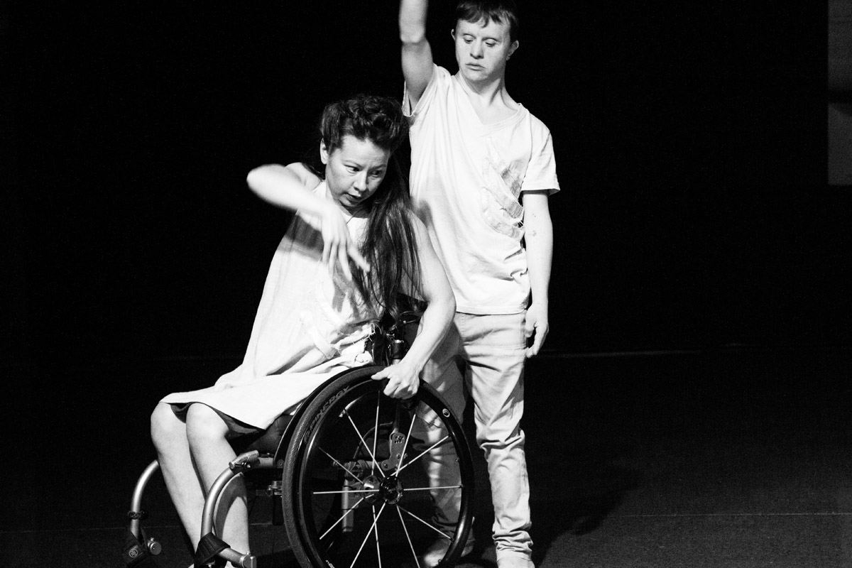 Two dancers perform on stage for Platform 12 Anniversary, one seated in wheelchair reaching across the body, the other standing behind reaching skyward.