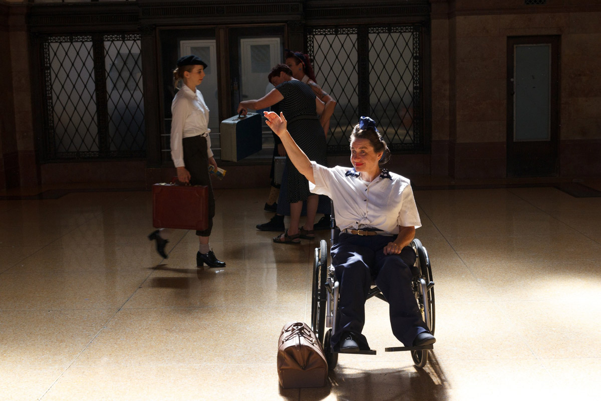 Four dancers on a dimly lit stage imitating a train platform. Centre stage dancer is seated in wheelchair while others stand in the background.